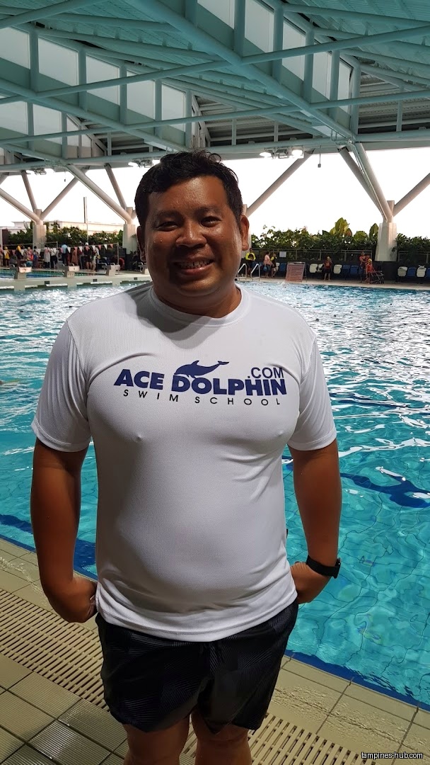 Man in Ace Dolphin Swim School shirt stands by a swimming pool.