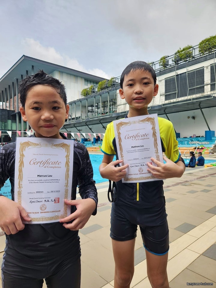Two boys in swimsuits hold certificates of completion near a pool.