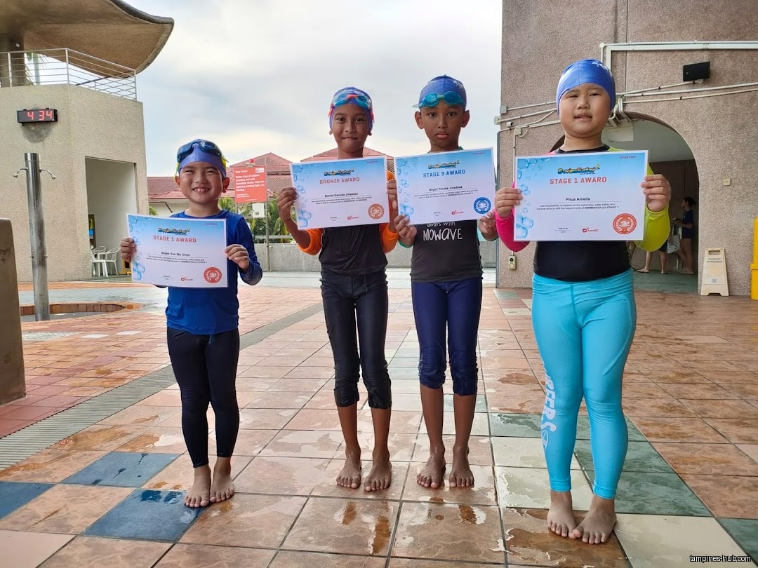 Four children in swimwear hold up swimming awards at a pool.