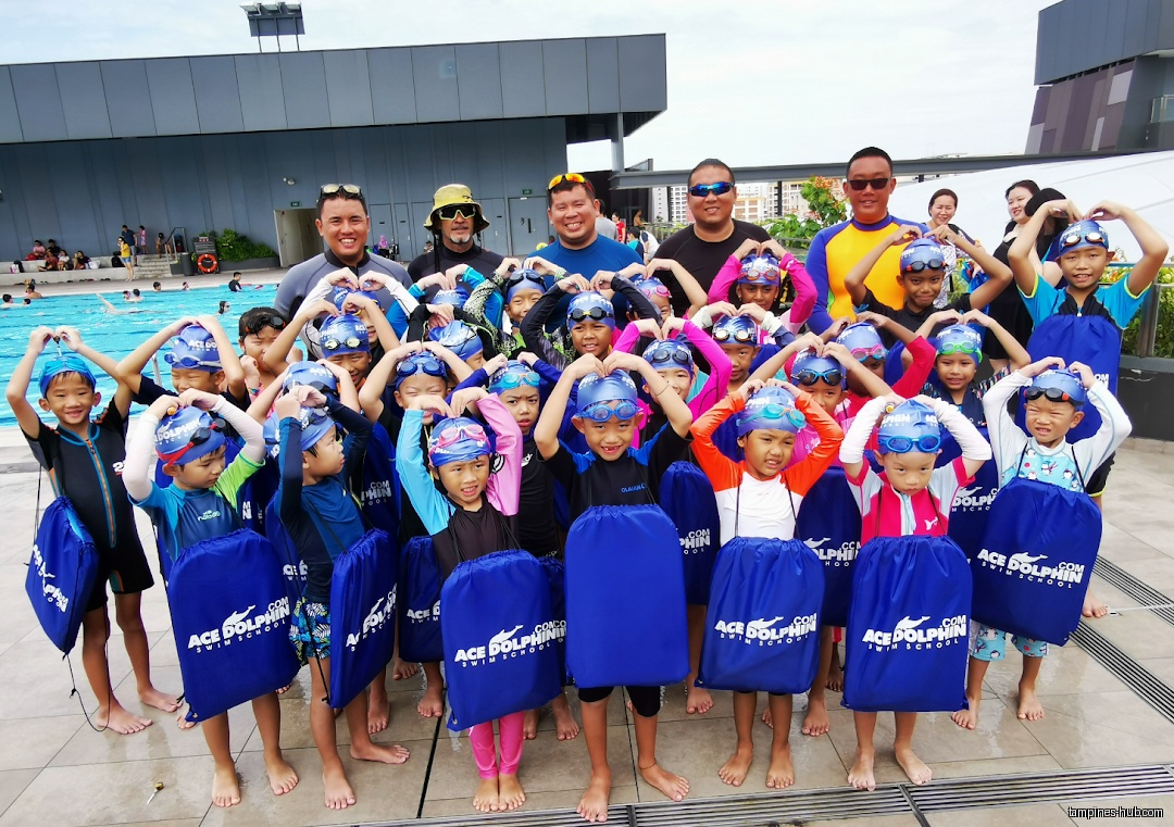 Group of children and adults pose with Ace Dolphin Swim School bags and swim caps.