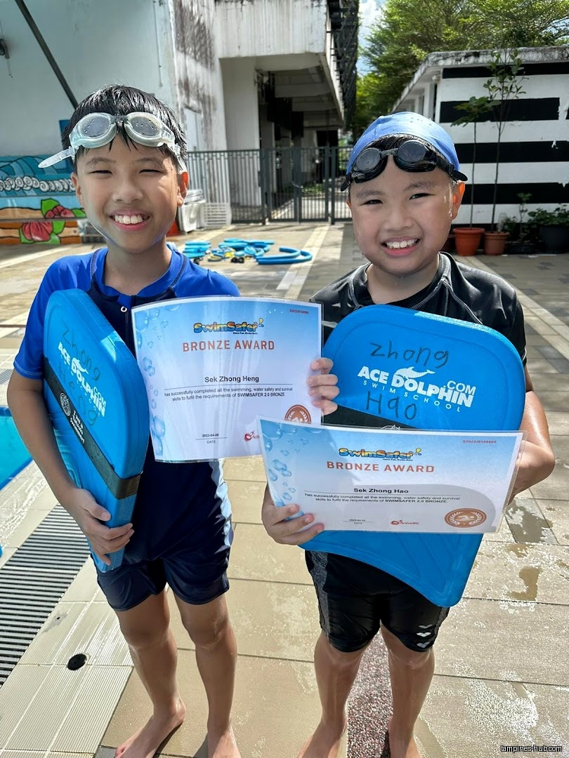 Two smiling boys hold swim boards and bronze awards at a pool.