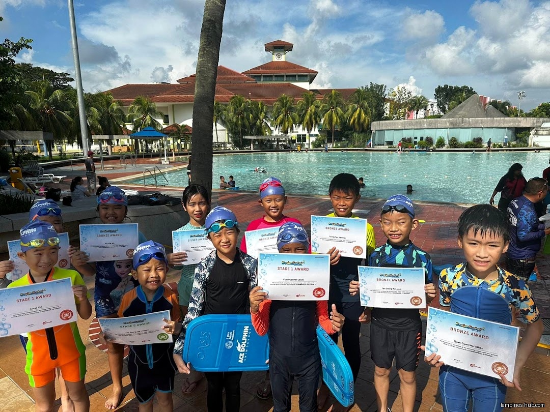 Children in swimwear hold swimming certificates near a pool at Ace Dolphin Swim School.