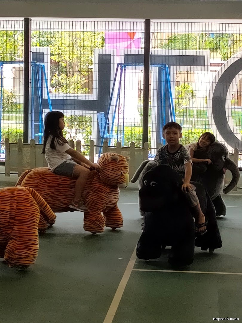 Children ride plush animal scooters in front of a window with shop signage.