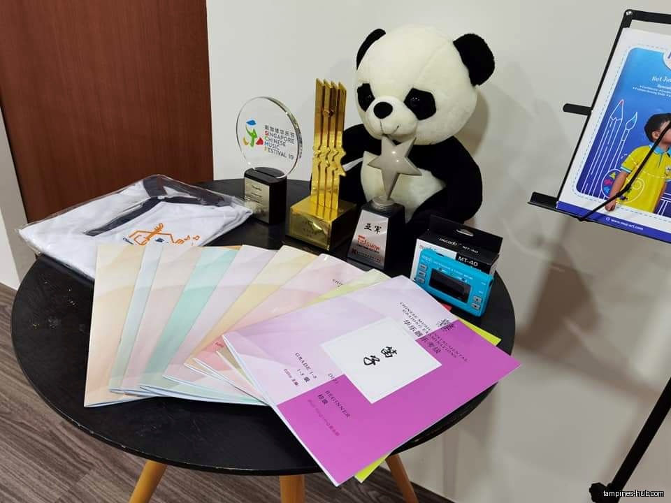 A table displays awards, a panda plush, music books, and a shirt.