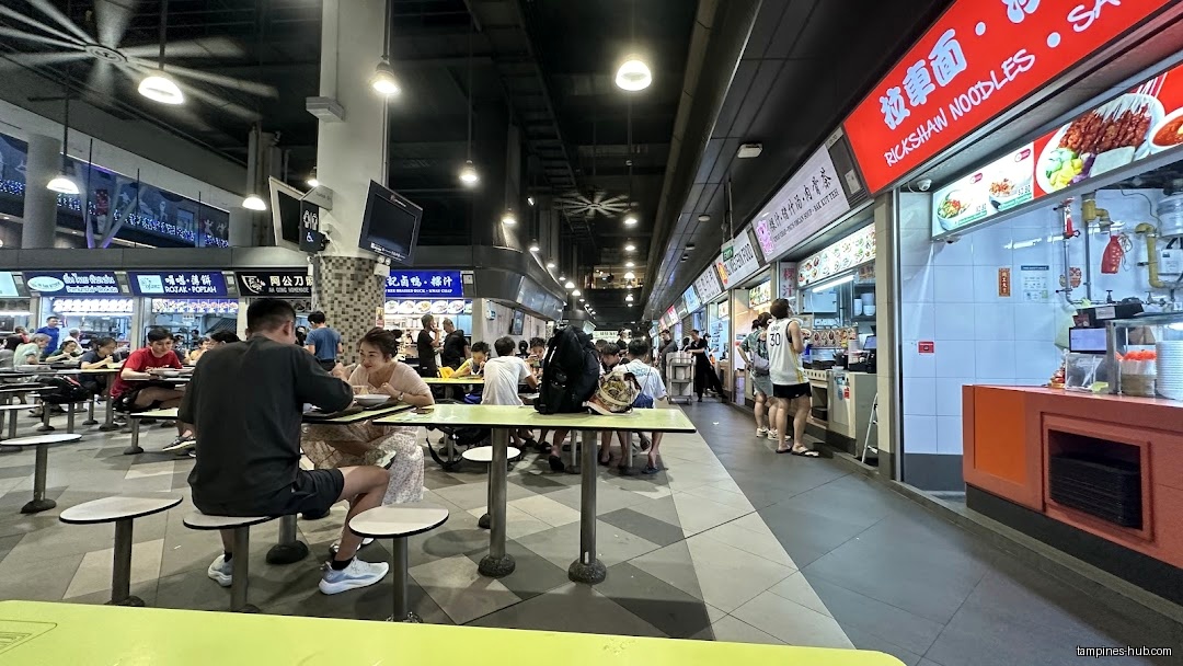 The image shows the interior of a busy hawker center with many people.