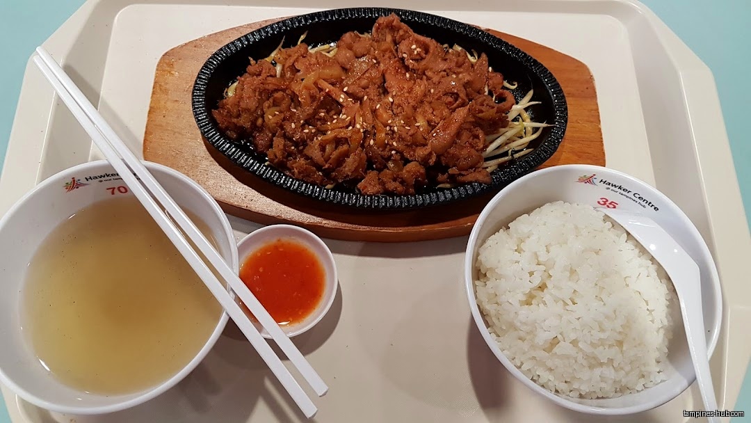 A tray with a sizzling plate of meat, rice, soup, and chili sauce.