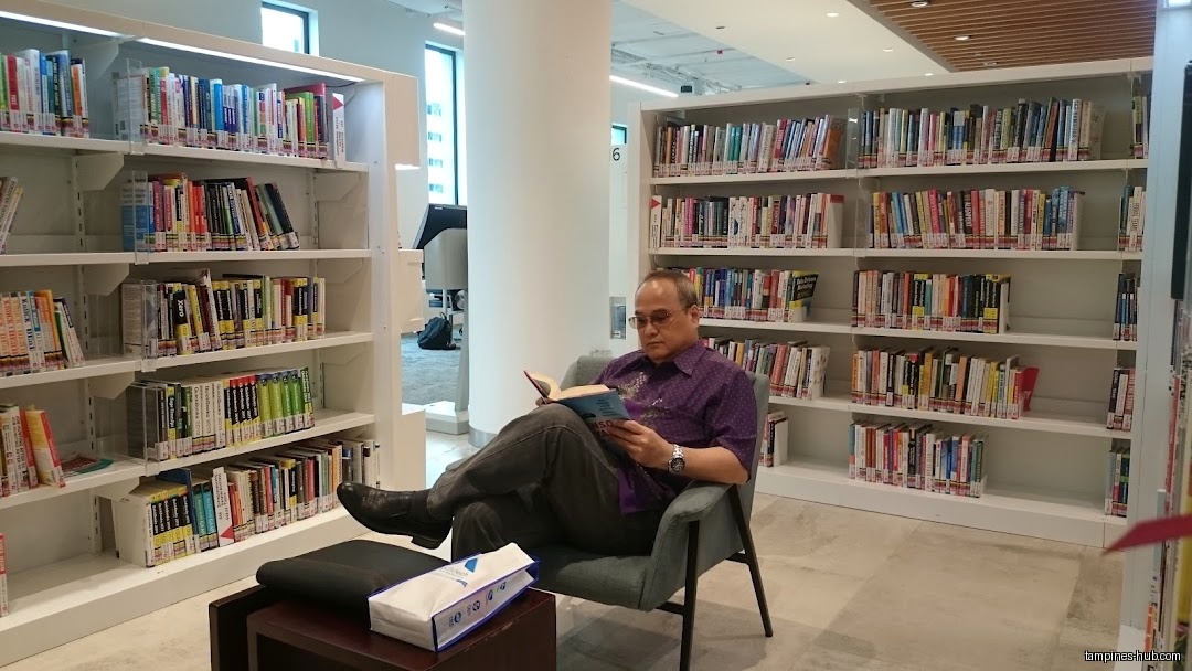 A man sits reading a book in a library with full bookshelves.