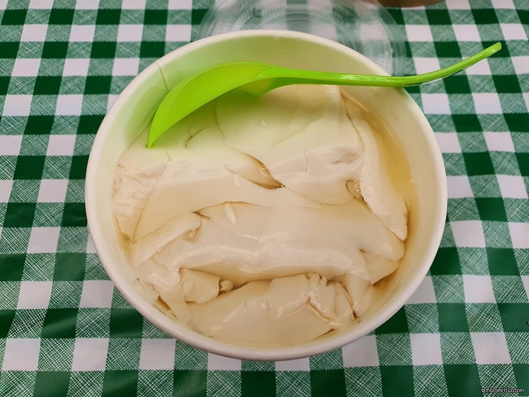 A bowl of tau fu fah with a green spoon on a checkered tablecloth.
