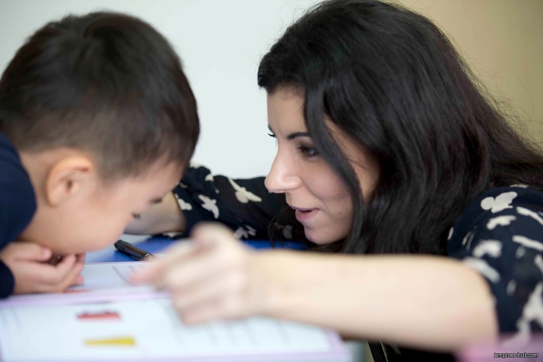 A teacher helps a young child with a worksheet at a desk.