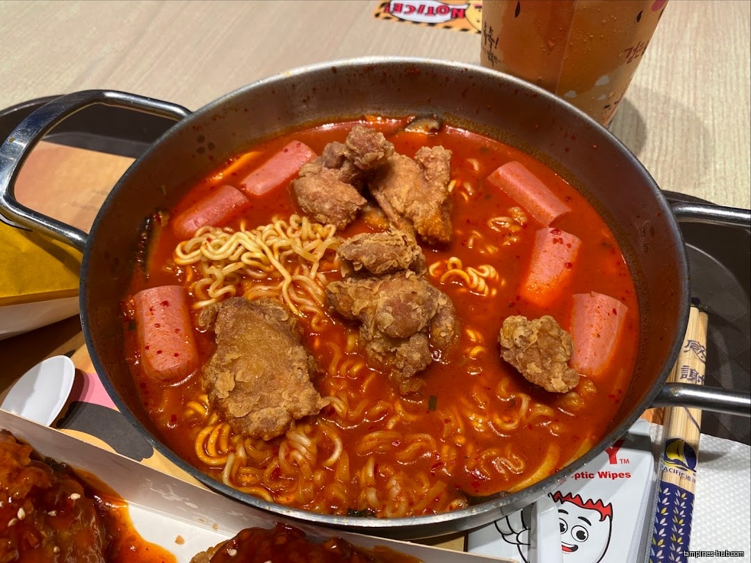 A pot of spicy ramen with fried chicken and sausage is shown.