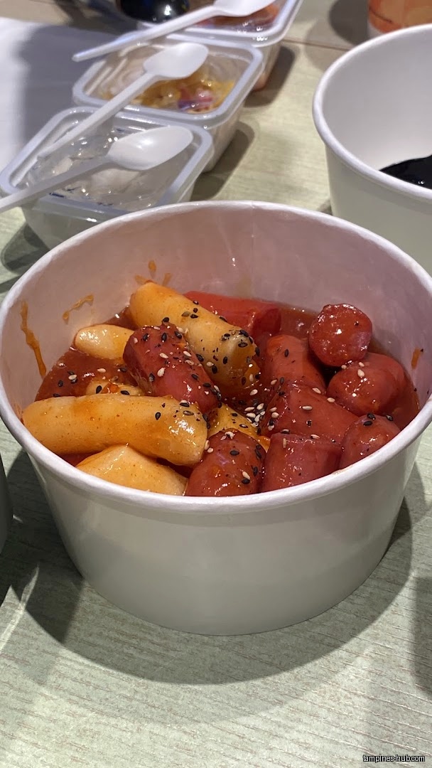 A bowl of tteokbokki with sausages and sesame seeds is shown.