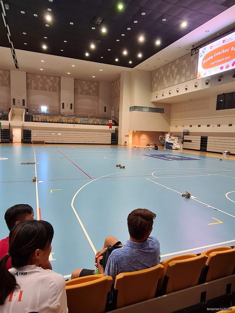 People sit in the stands overlooking a basketball court in a large auditorium.