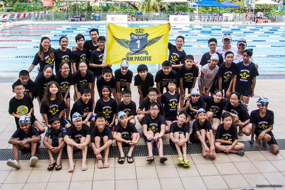 A large group of people wearing Pacific Swim Team shirts pose by a pool.