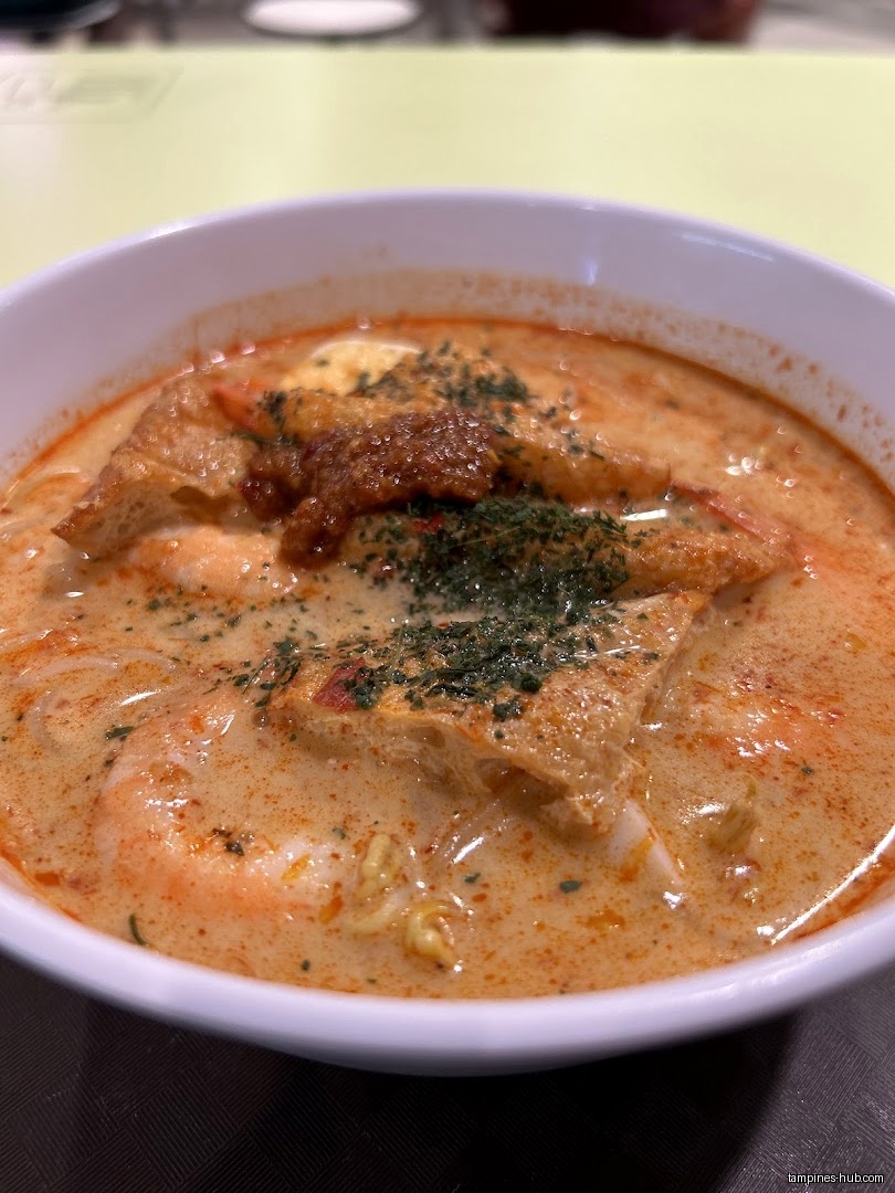 A bowl of laksa with tofu, shrimp, and herbs is shown.