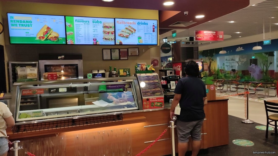 A Subway restaurant interior with menu boards and a customer waiting.