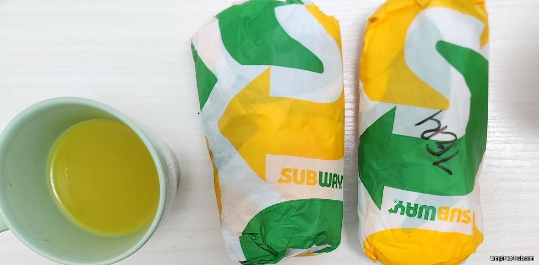 Two Subway sandwiches in wrappers and a cup of yellow liquid.