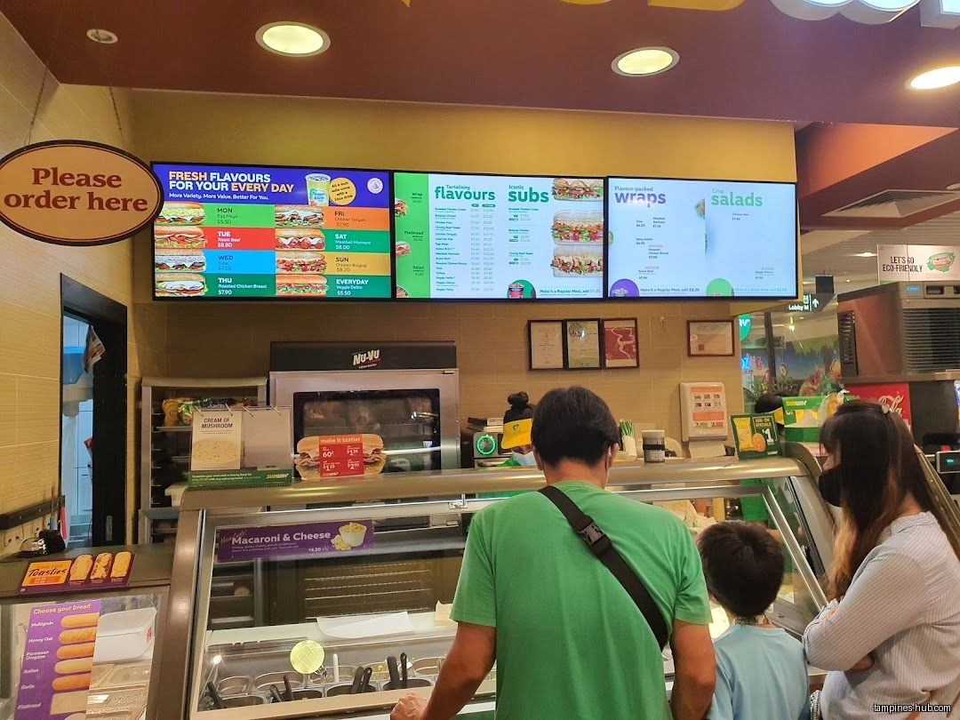 Customers are ordering at a Subway restaurant, viewing the menu boards.
