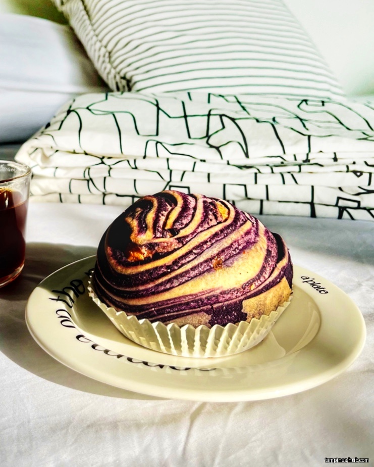 A purple swirl bun on a plate with coffee and bedding in background.