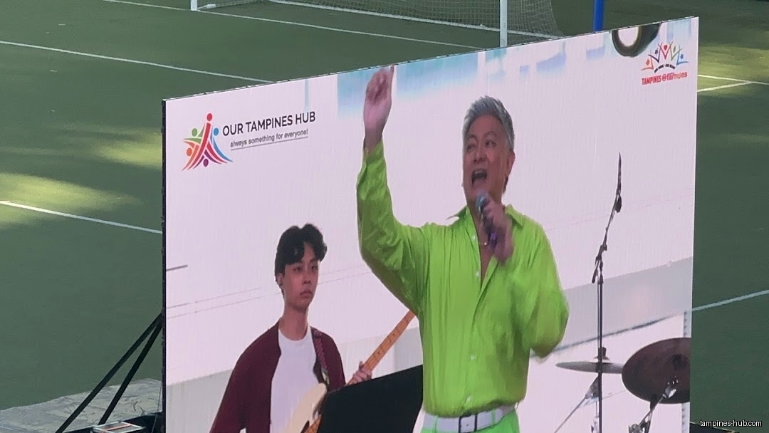 A man in green sings on a screen with a band behind him.