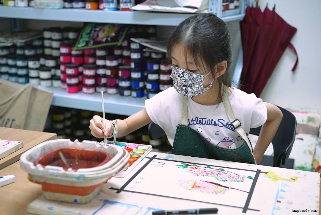 A young girl wearing a mask paints at a table in an art shop.