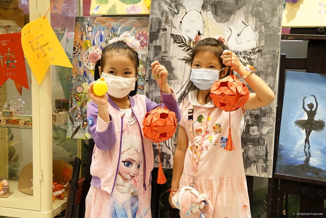 Two girls in masks hold lanterns in front of artwork at Tree Art.