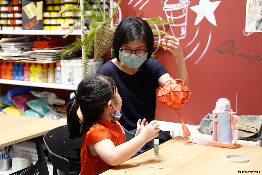 Child and adult making an orange paper lantern at a table.