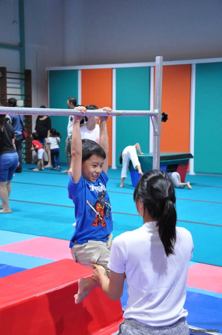 A child hangs from a bar with an instructor nearby in a gym.