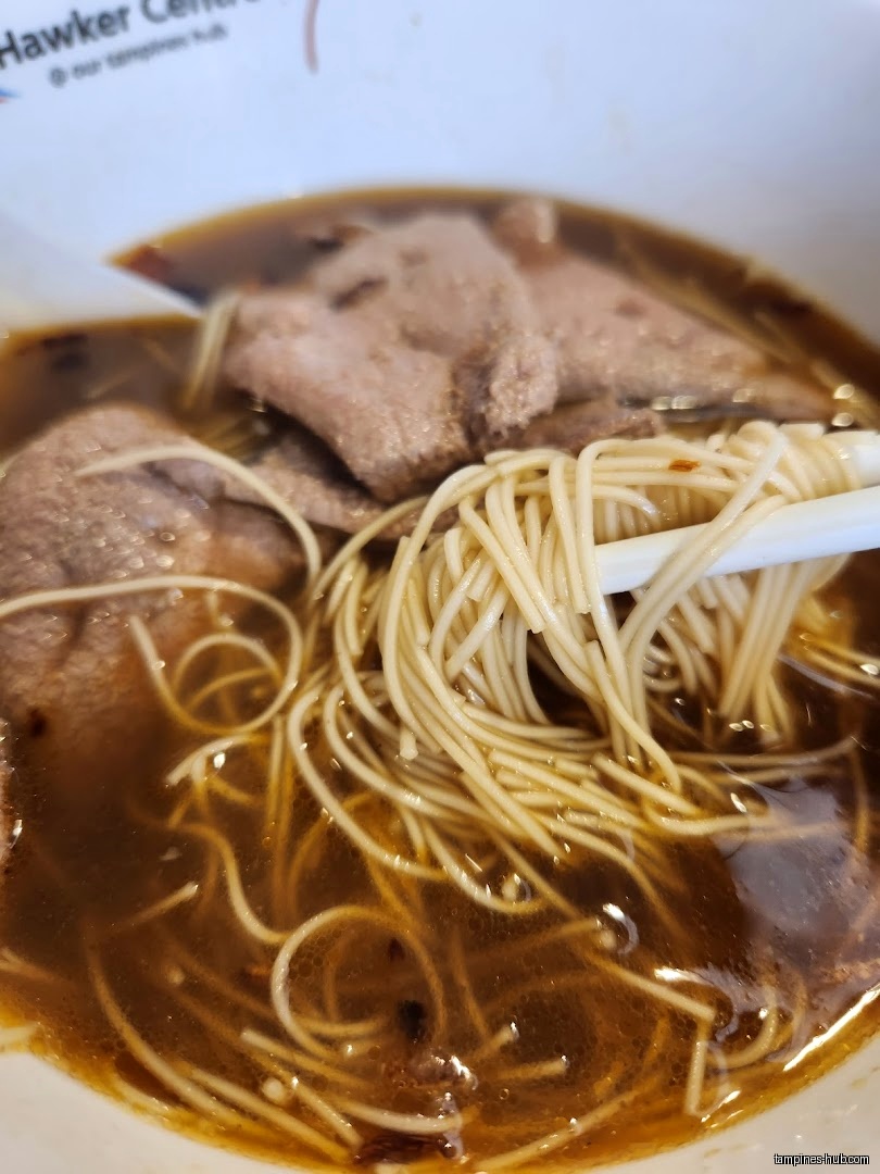 Close-up of prawn noodles with broth and meat, held by chopsticks.