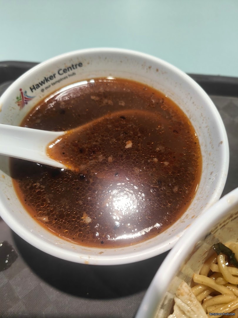 A bowl of dark soup with a spoon, next to noodles.
