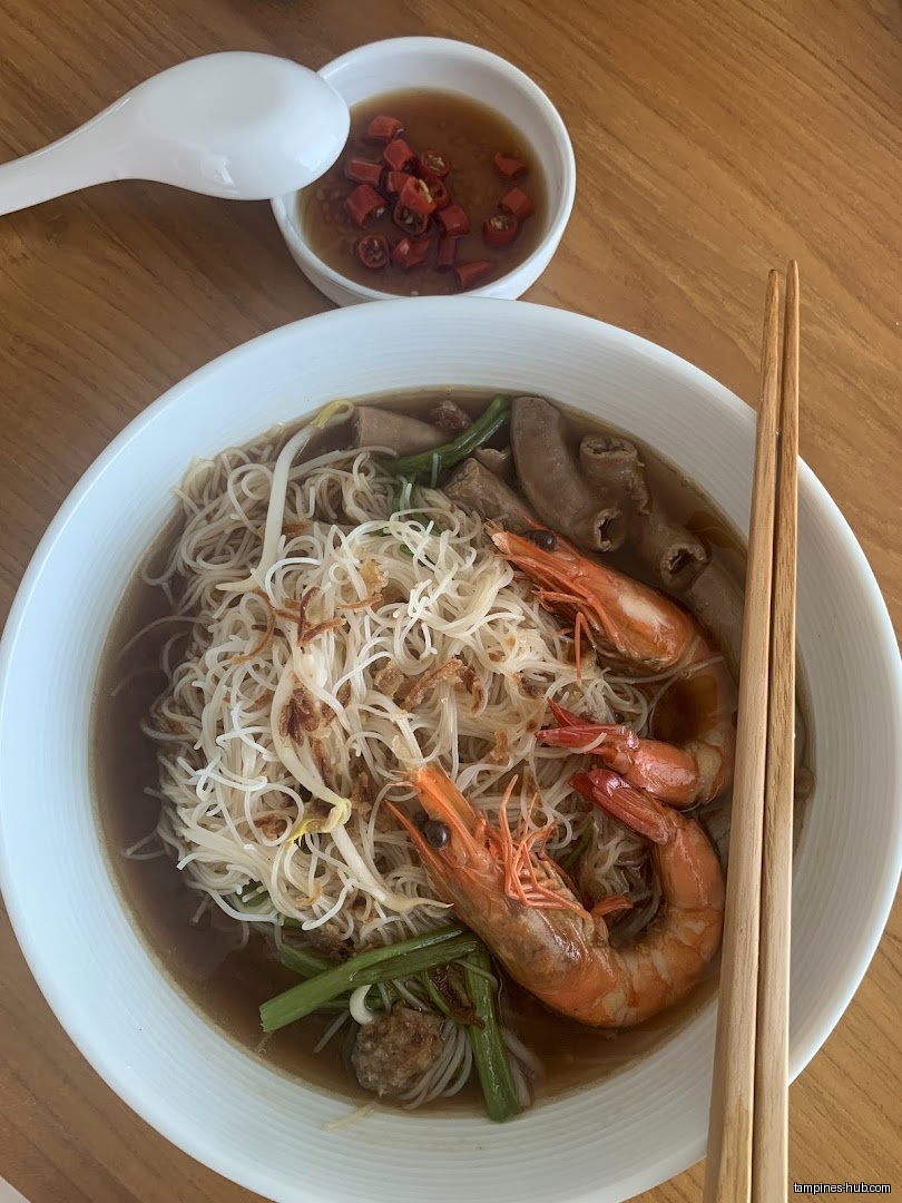 A bowl of prawn noodles with prawns, vegetables, and a side of chili.
